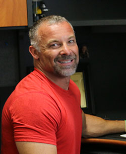 Gary Ackerman Plant Manager stitting at Desk in orange shirt smiling at work