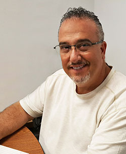 Greg Collins President and Sales sitting at desk in a white shirt with glasses smiling at work
